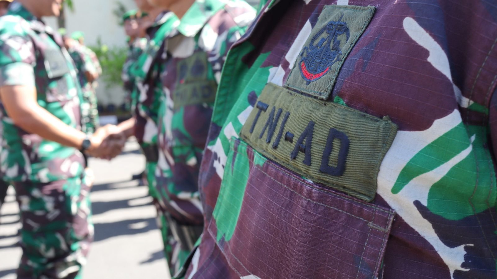 Seragam loreng TNI Angkatan Darat, dengan prajurit berjabat tangan di latar belakang